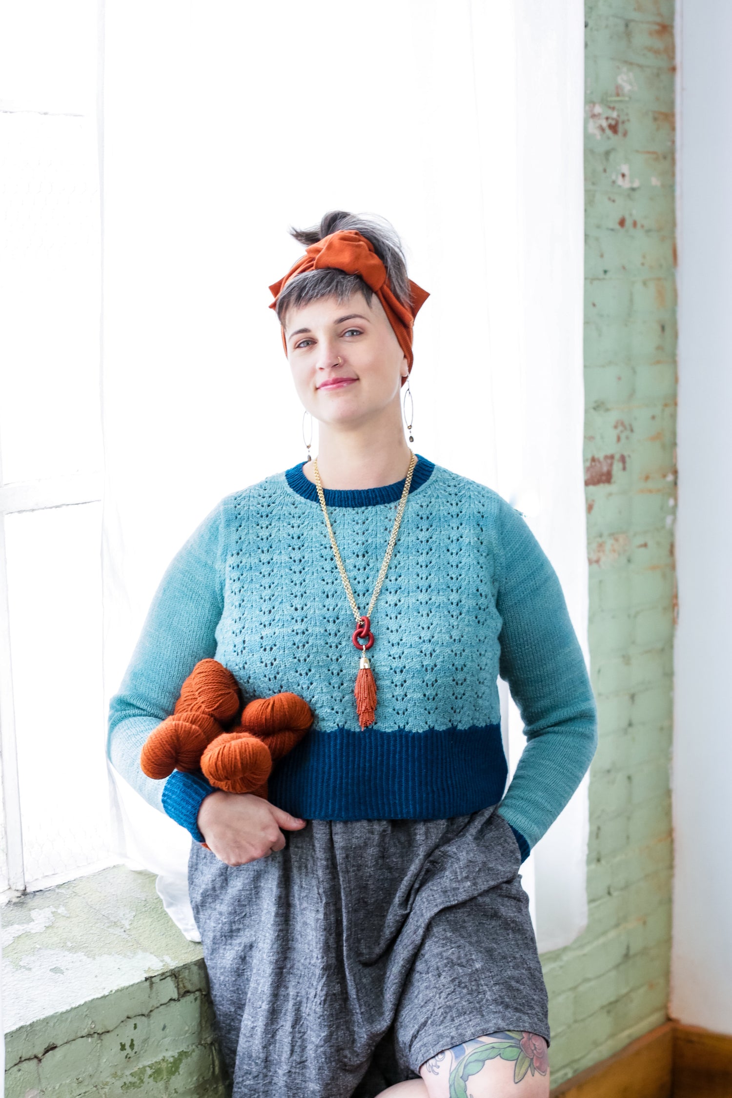 Jen leans against a window ledge, holding four skeins of yarn and smiling at the camera. She wears a light and dark blue lace knit sweater, knit from her Classic Colorblock pattern.