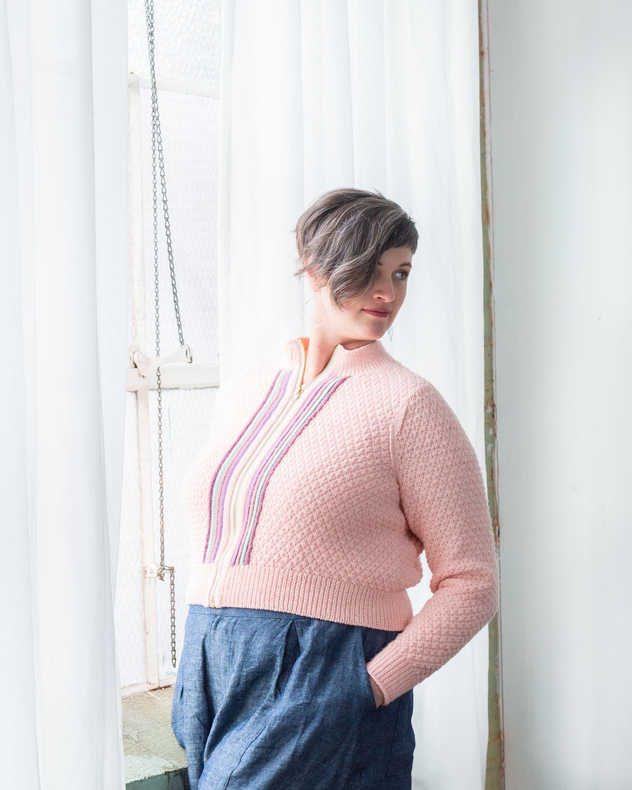 Jen, leaning against a window frame and looking off camera, wears a blue skirt with a hand knit, light pink jacket. The jacket features four plackets around a gold zipper, with a high collar.