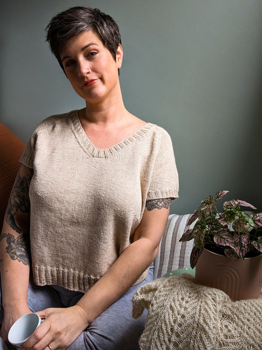 Jen sits on a couch, smiling at the camera. She wears a light beige version of her Classic LBD pattern, knit with short sleeves. A knit blanket and potted plant sit next to her.
