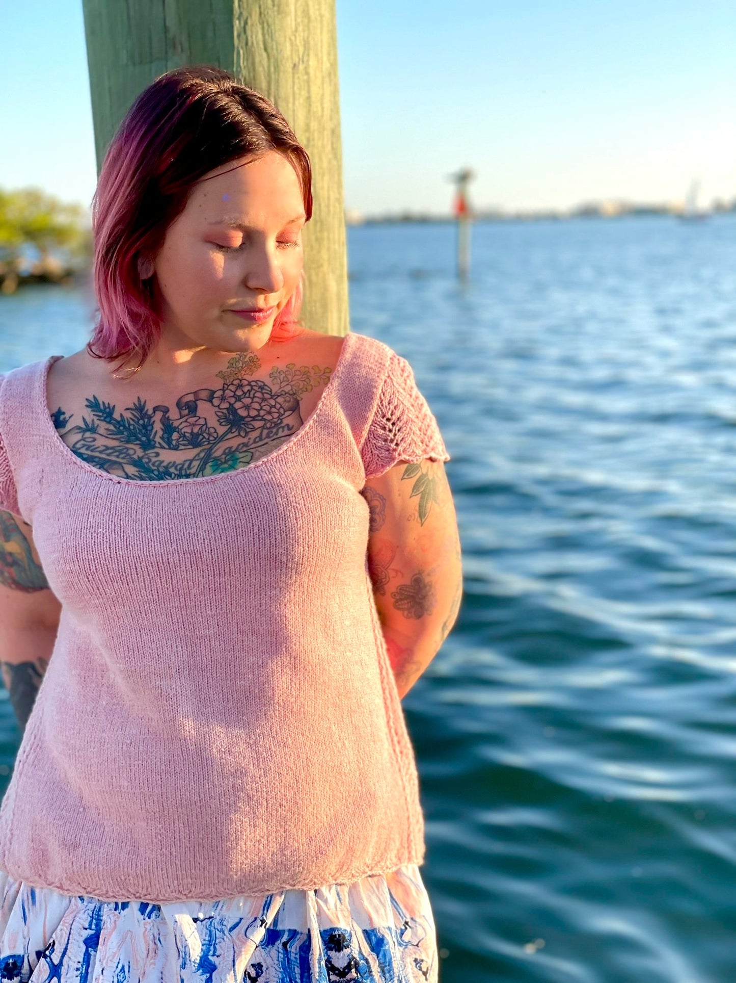 A woman wears a light pink wool tee with a gentle a-line shape over a ruffled white printed skirt. The tee is knit from a knitting pattern featuring lace cap sleeves and lace inserts along each side.