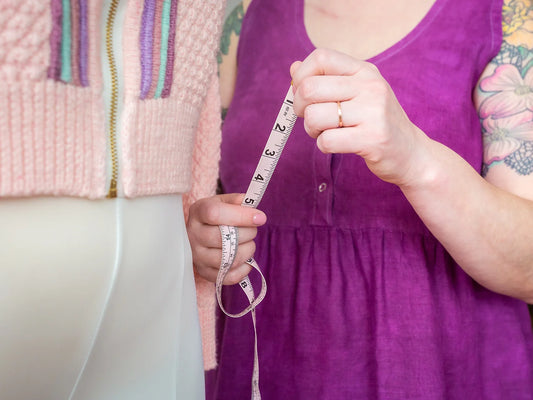 Jen, seen from the neck down, holds a dressmaker's tape up to measure the Letters from the Open Road sample on a dress form. She wears a magenta tank dress. The sample sweater is knit with light pink yarn and multicolored plackets around the zipper.