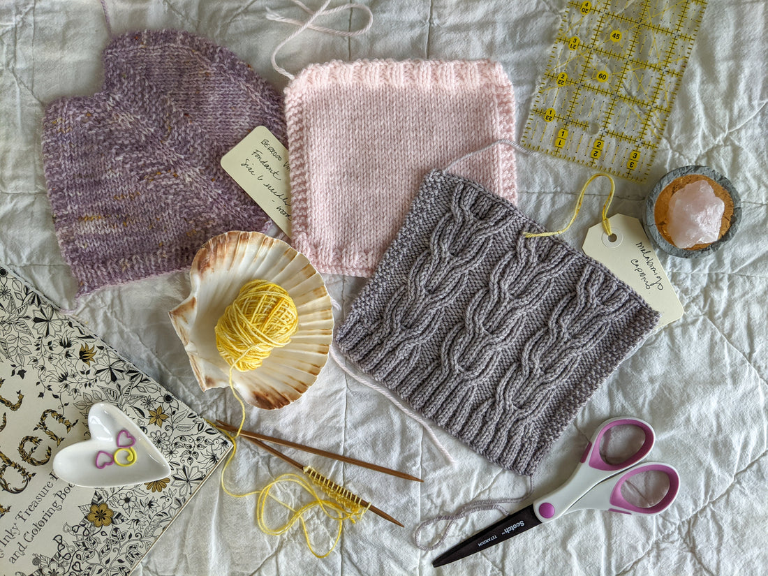 Three gauge swatches, one in purple stockinette, one in light pink stockinette and rib, and one in a grey cable design, lay on a tabletop next to small balls of yarn, scissors, and stitch markers.