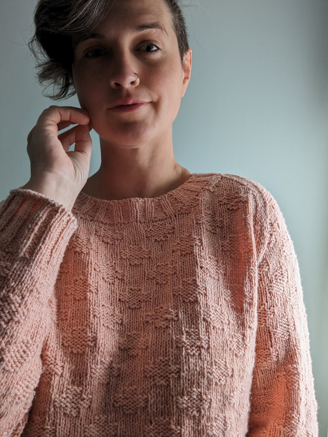 Jen looks at the camera, wearing a coral pink version of her Plusses sweater, knit using a textured plusses design instead of the colorwork pattern.