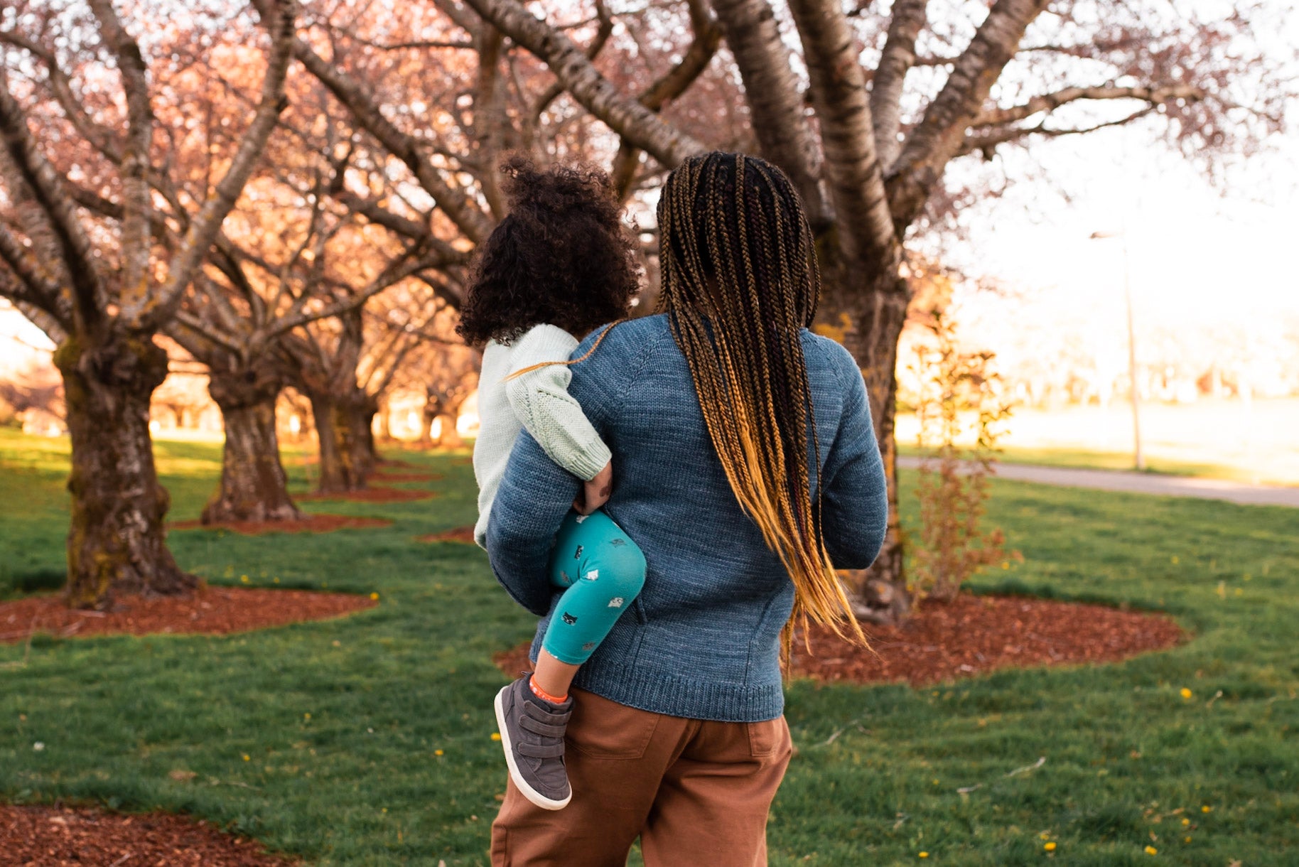 Seen from behind, Rachel carries Mia through a park. They wear matching hand knit sweaters with subtle cable detailing. Rachel's sweater is knit in blue yarn, and Mia's in cream.