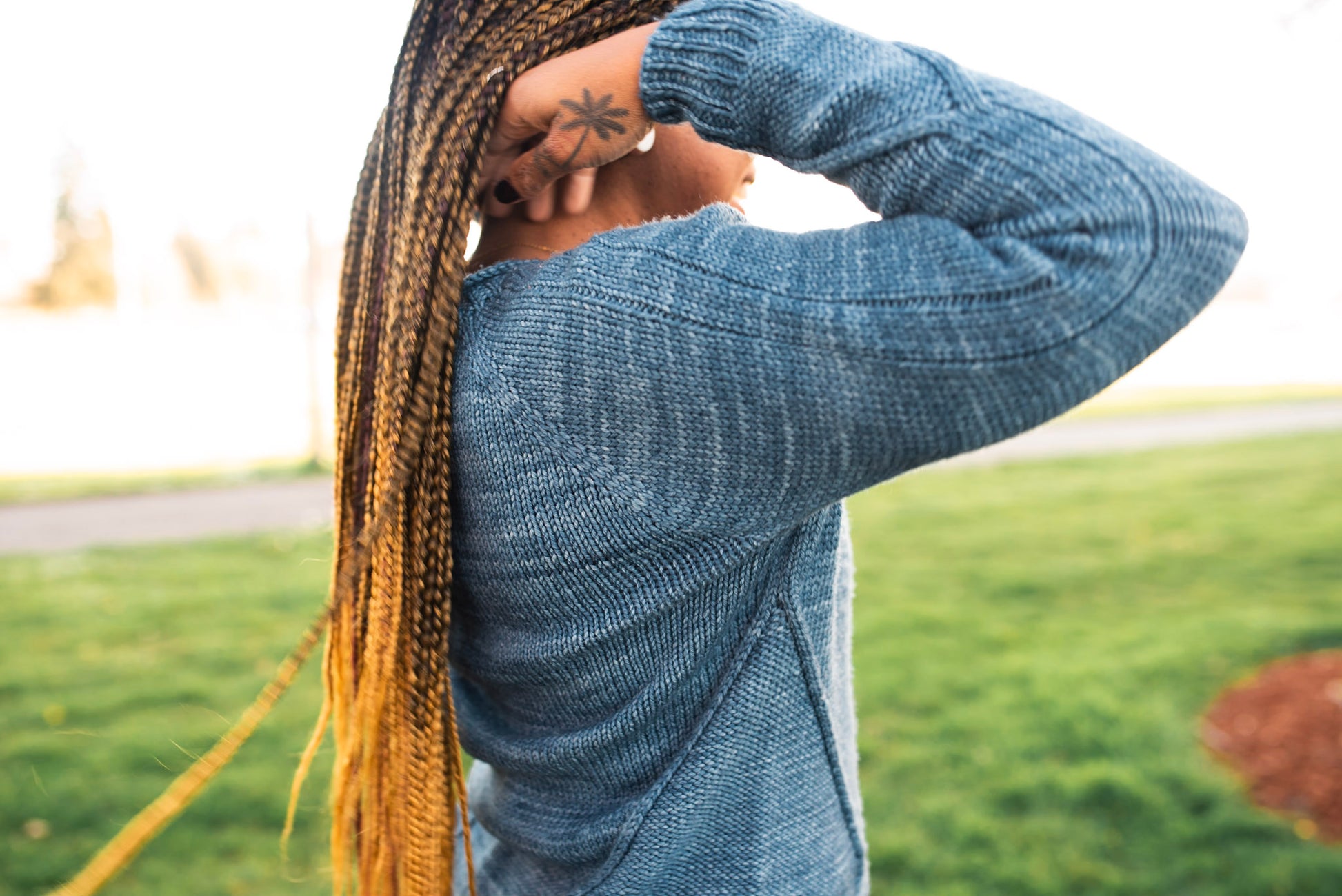 Seen from behind at a three quarter angle, Mia smiles while moving her braids off her shoulders. The camera focuses on the cable details of her hand knit blue sweater.