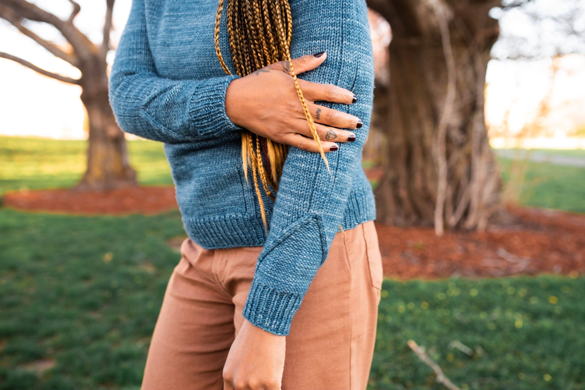 Seen from the shoulder's down, Rachel stands in a park wearing a hand knit blue sweater with pink pants. The sweater has subtle cable details around the cuffs.