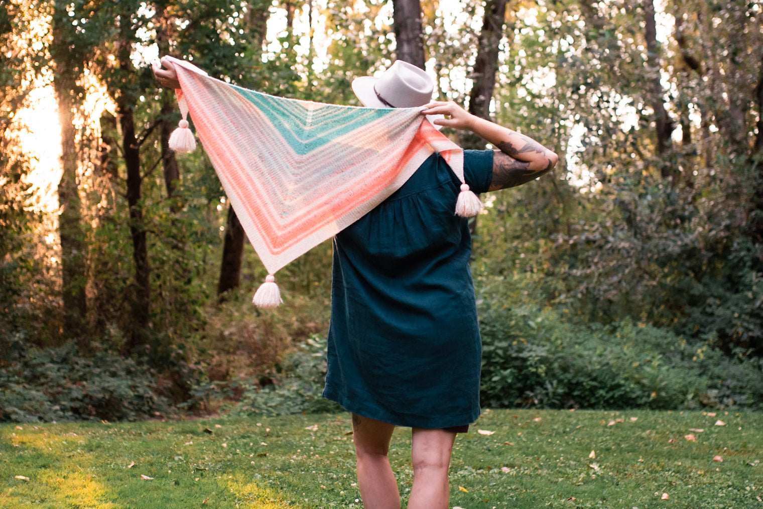 Seen from behind, Jen holds a pastel striped shawl, knit in a triangle shape with tassels at each corner. She wears a white hat with a blue dress. Trees can be seen in the background. 