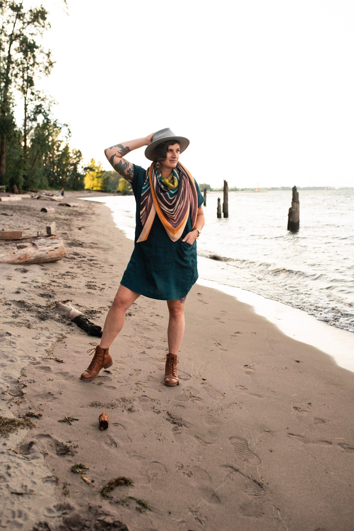 Jen stands on a beach, looking off camera and wearing a hand knit striped shawl with a blue dress and a white hat. 