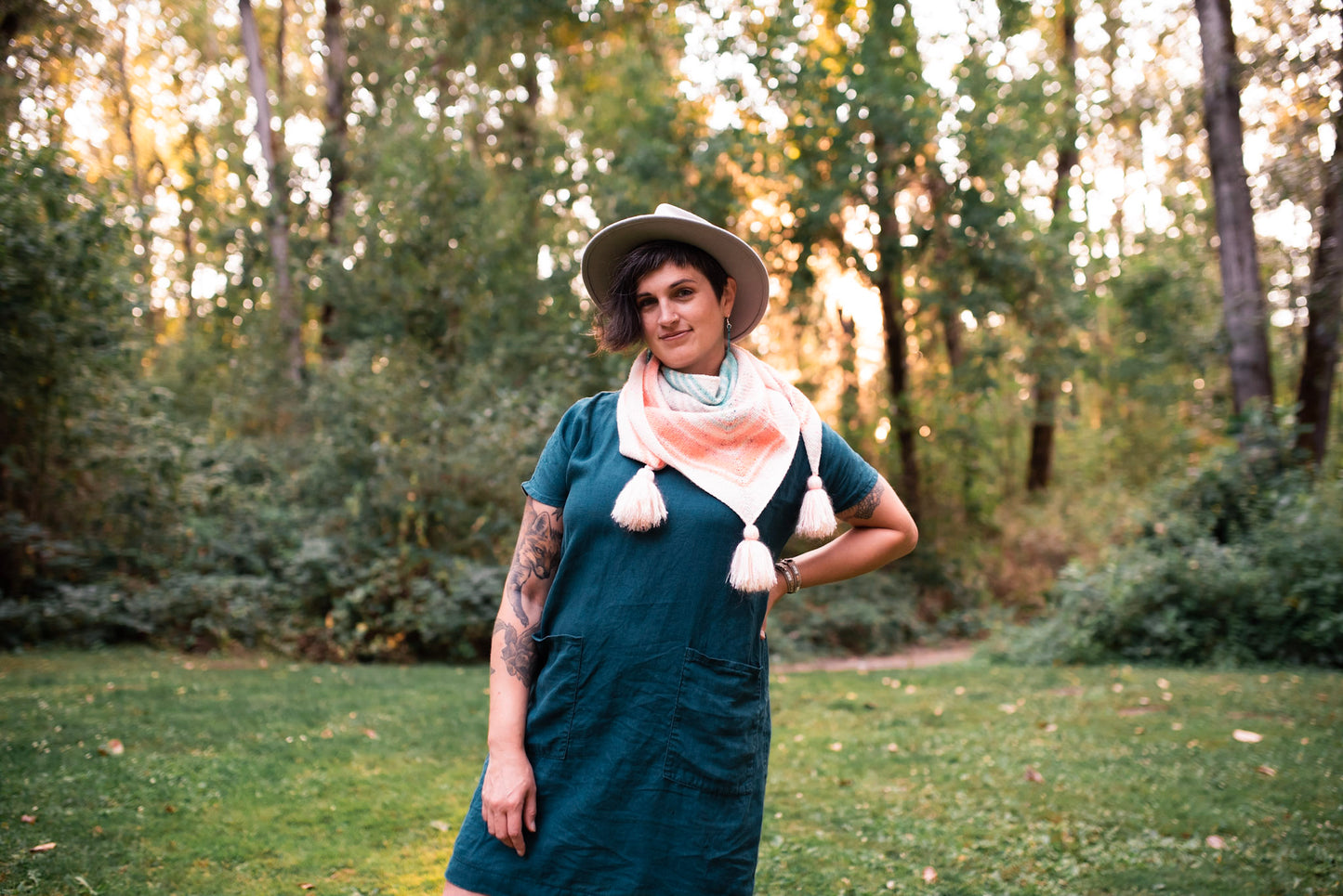 Standing in a park, Jen wears a pastel knit shawl with a blue dress and a white hat. The shawl has tassels at each corner. Trees can be seen in the background.