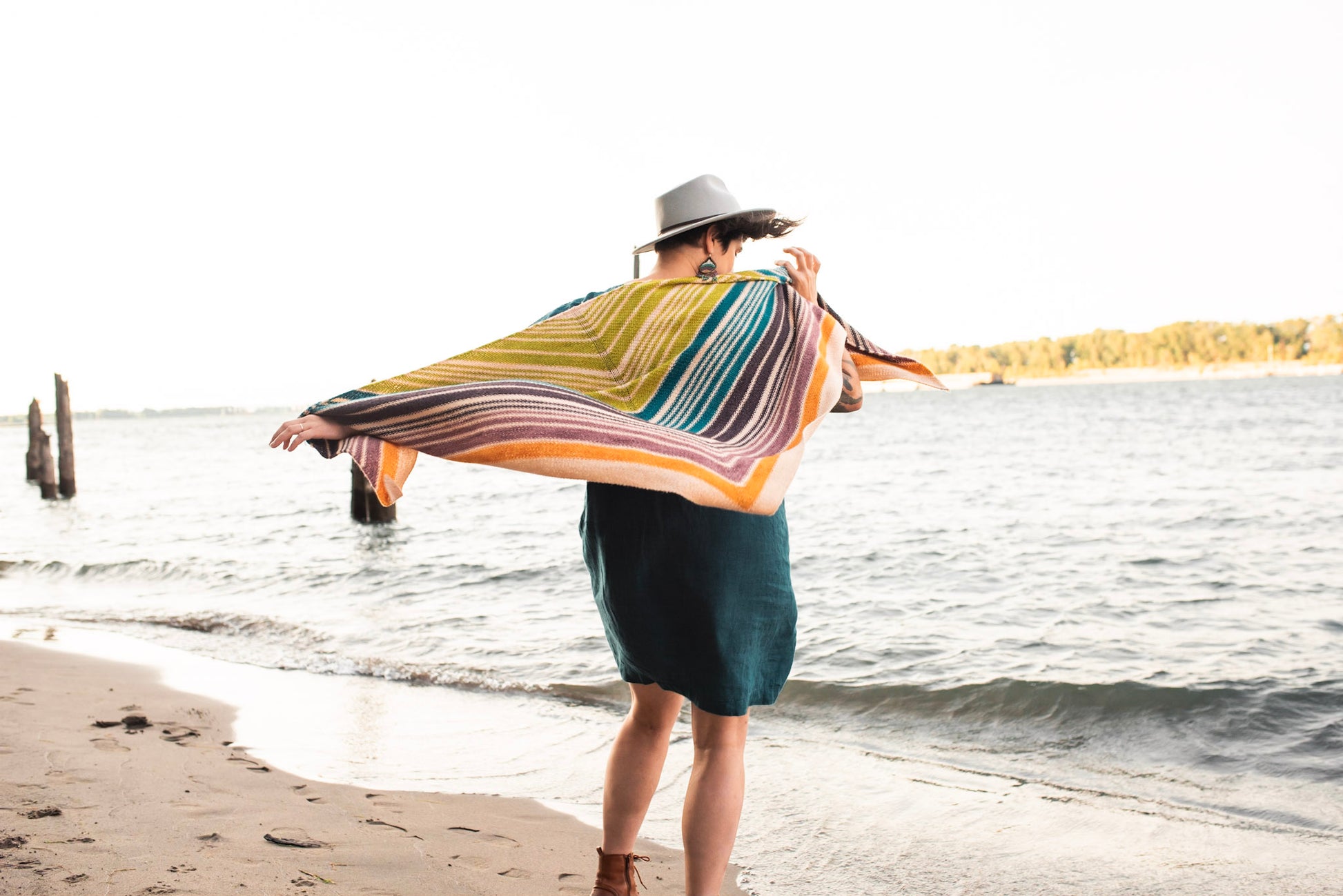 Seen from behind, Jen wears a striped shawl with a blue dress and a white hat. The shawl is knit in a triangle shape, and flows in the wind. A beach and waterfront can be seen in the background.