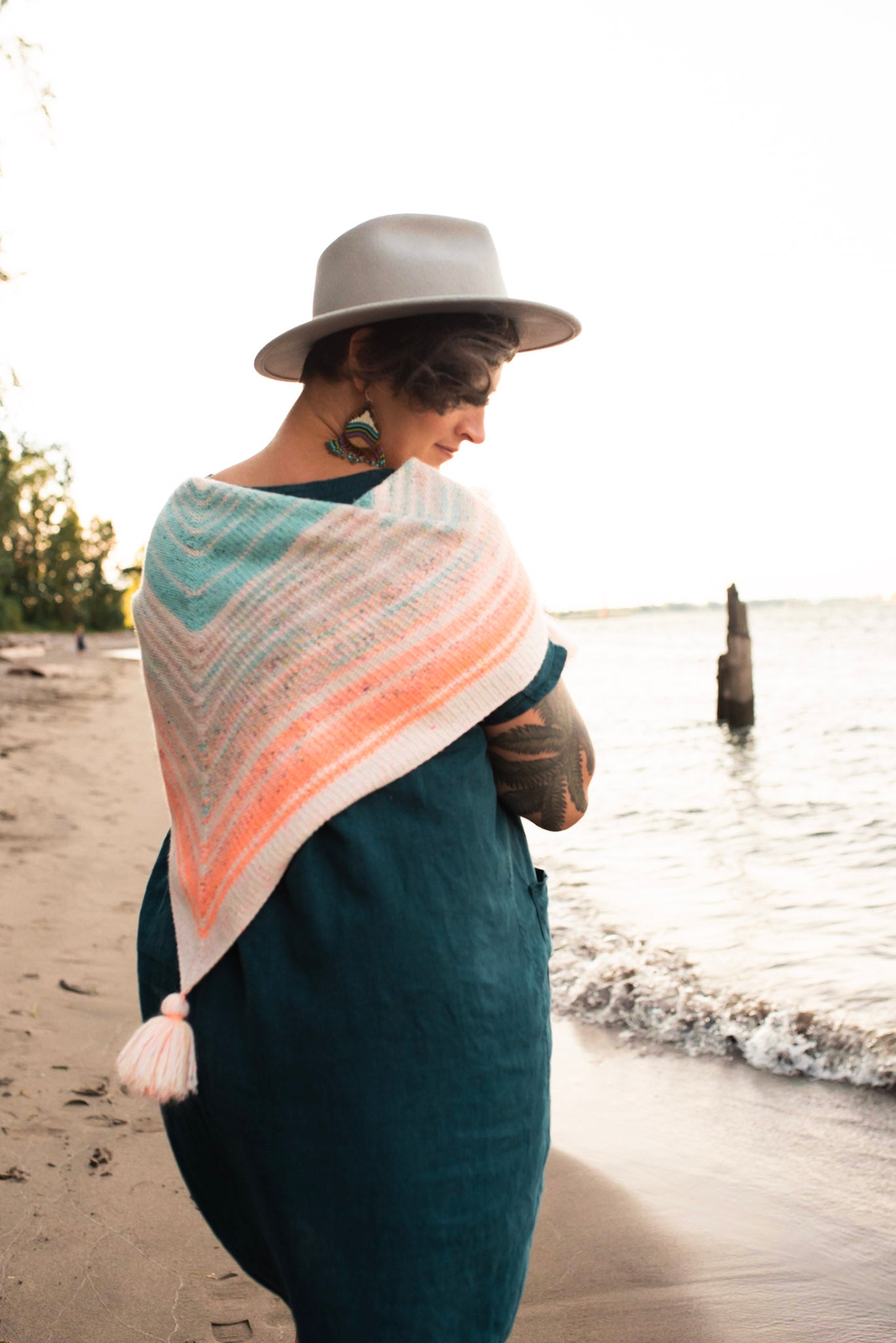 Seen from behind, Jen wears a pastel striped shawl, draped over her shoulders. The shawl has tassels at each point of the triangle shape. A beach and some trees can be seen in the background.