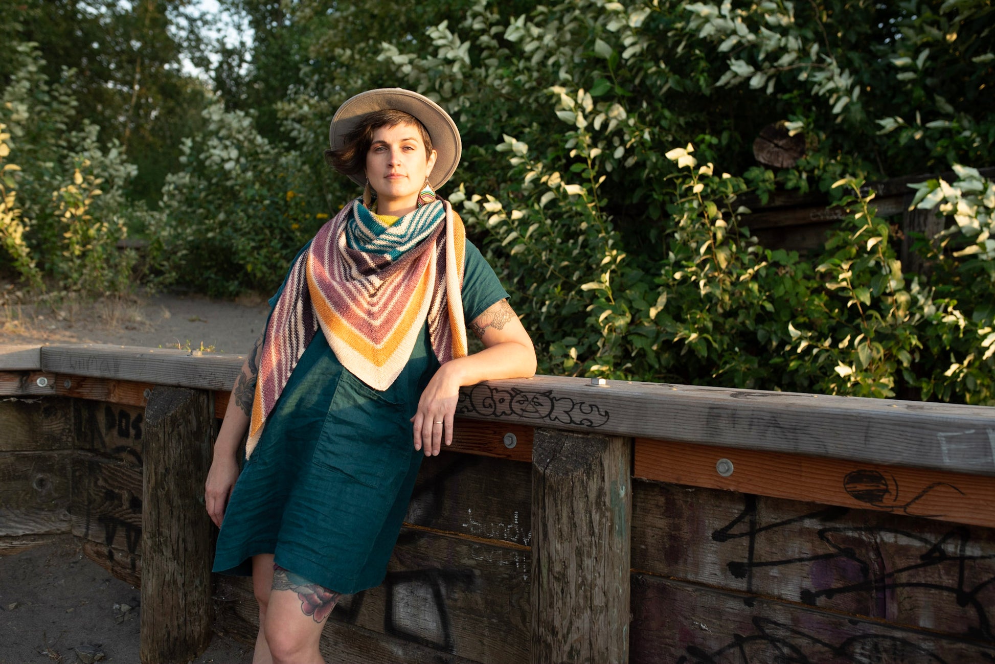 Jen leans against a wooden railing, smiling at the camera. She wears a multicolored shawl, knit in a triangle shape, with a blue dress and a white hat.