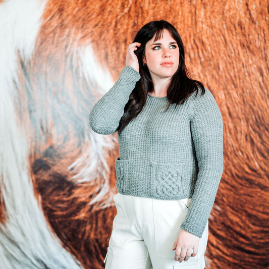 Becca, standing in front of a large photo, wears white pants with her hand knit sweater. The sweater features two pockets with a cable design on them. 