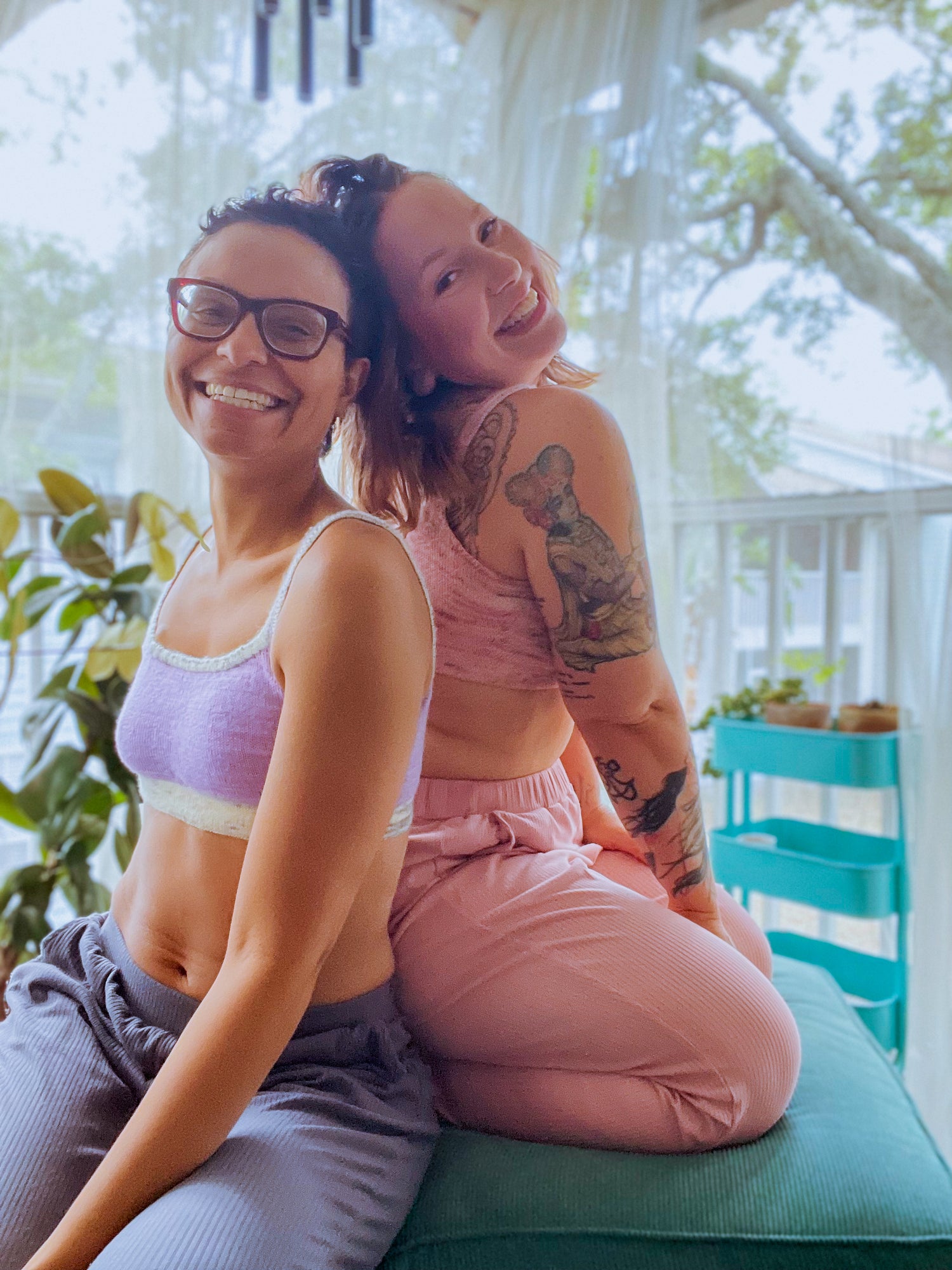 Bess and Candace sit on a sofa footstool, grinning at the camera. Bess wears a light pink version of the Carol Bralette with matching sweatpants. Candace wears a light purple bralette, knit with a white speckled band and straps, with matching purple pants.