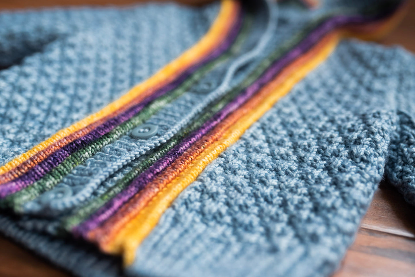 Seen close up, a light blue moss stitch kid's sweater lays on a table. It has four blue buttons and yellow, orange, purple, and green plackets around the button band.