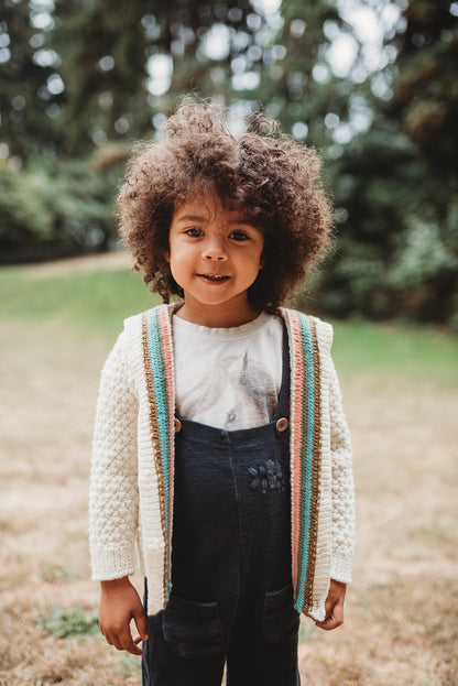 A young boy stands in a park, wearing black overalls and a white shirt under a hand knit sweater. The sweater is unbuttoned and knit with cream yarn, featuring orange, green, brown, and blue plackets around the button band.