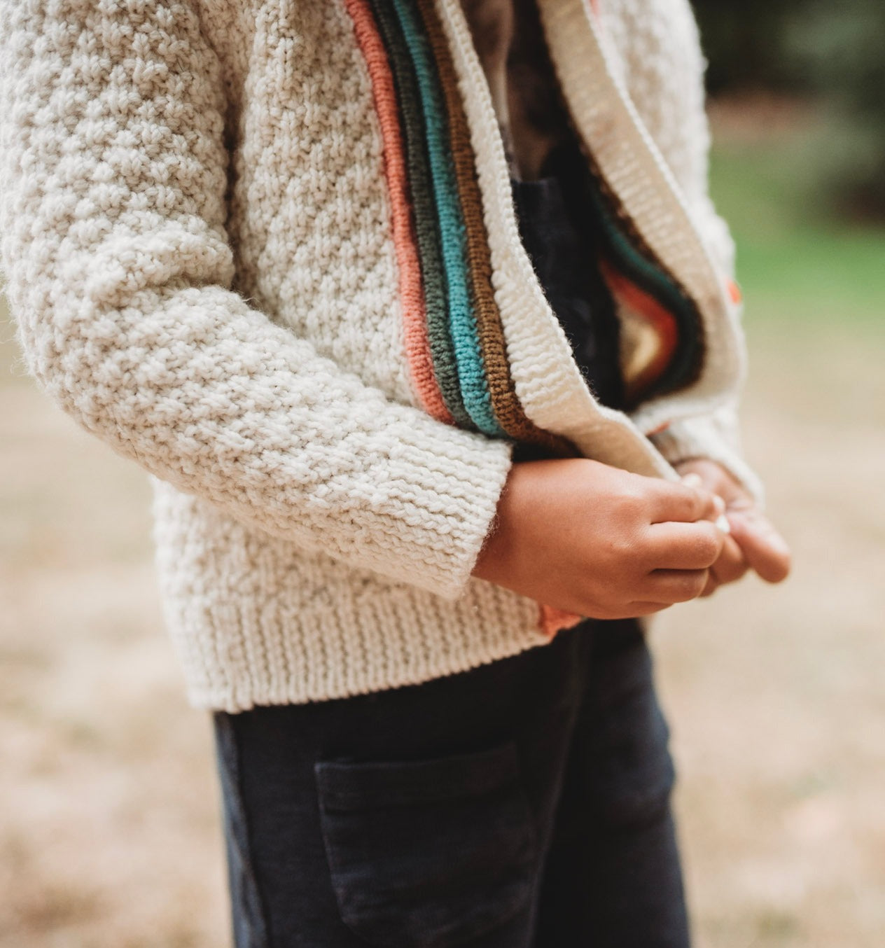 Seen close up from a 3/4 angle, a young boy buttons up hand knit jacket, knit in cream with brown, green, blue, and orange plackets around the button band.
