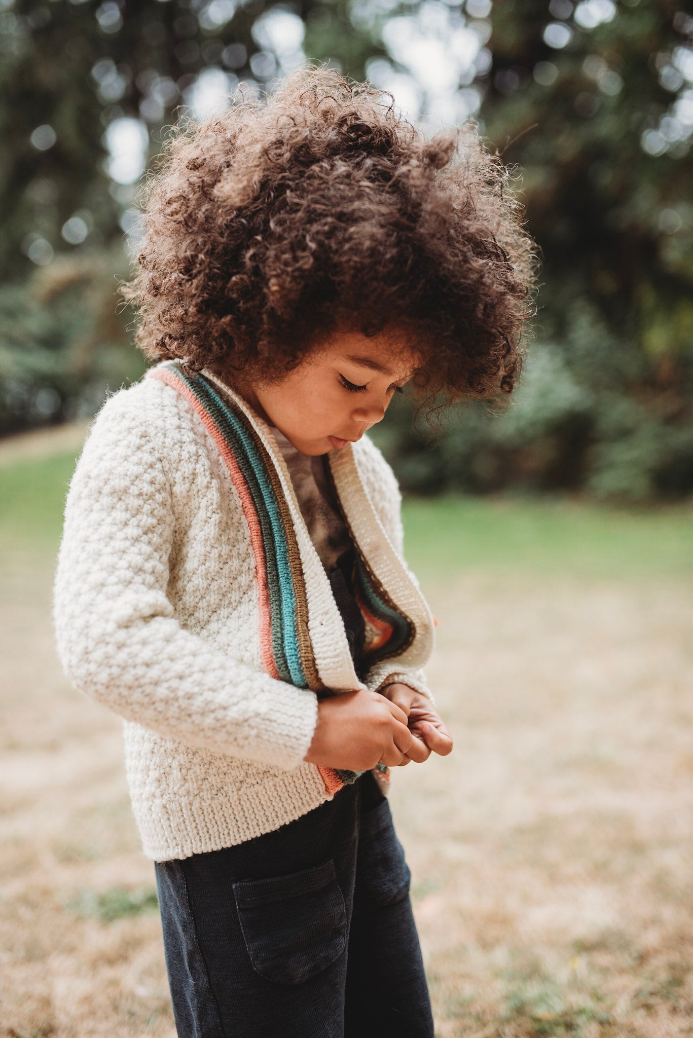 A young boy with curly brown hair, seen from a 3/4 angle, buttons up hand knit jacket, knit in cream with brown, green, blue, and orange plackets around the button band.
