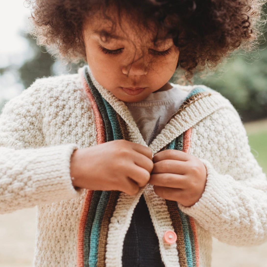 A young boy, seen close up, focuses on buttoning up his cream knit sweater. The sweater features orange, green, blue, and brown plackets around the cream button band.