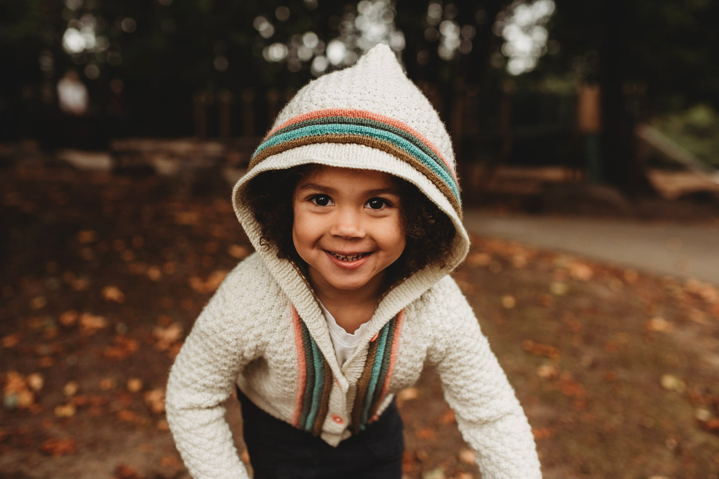 A young boy smiles at the camera and wears a Letters to Camp sweater, buttoned up and with the hood up. It's knit with cream yarn in a moss stitch, with orange, green, blue, and brown plackets around the button band. A park can be seen in the background.