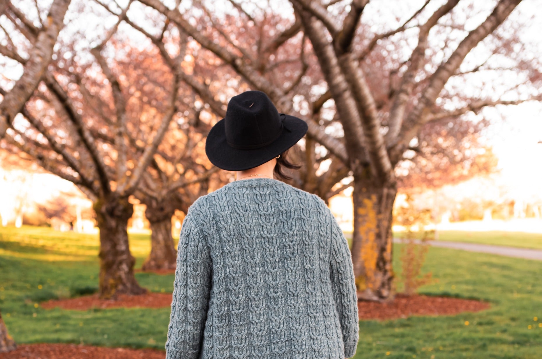 Seen from behind, Jen wears a black hat with a cable knit cardigan. A park with a grove of trees can be seen in the background.