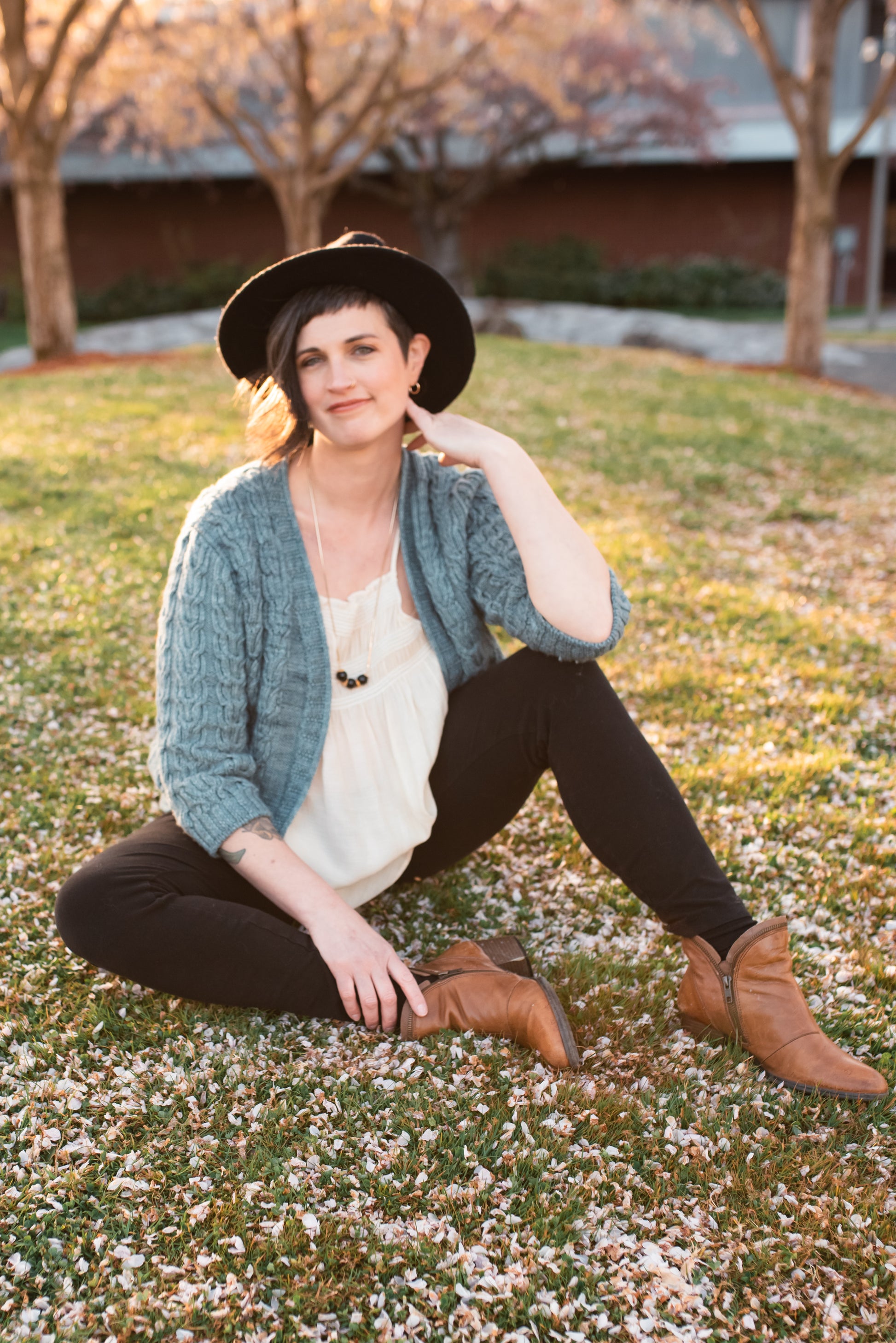 Sitting on the ground, Jen smiles at the camera, wearing a light blue cardigan knit with three quarter length sleeves over a white tank top.