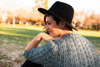 Seen from behind, Jen wears a black hat with a hand knit, light blue cardigan, adorned with horseshoe cables and a ribbed neckline.