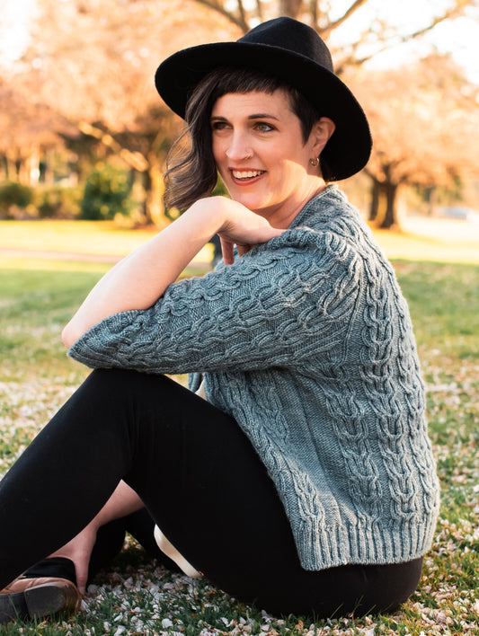 Jen sits in a park, seen from the side. She wears a black hat and black pants with a light blue cardigan. The cardigan is knit with horseshoe style cables and 3/4 length sleeves.