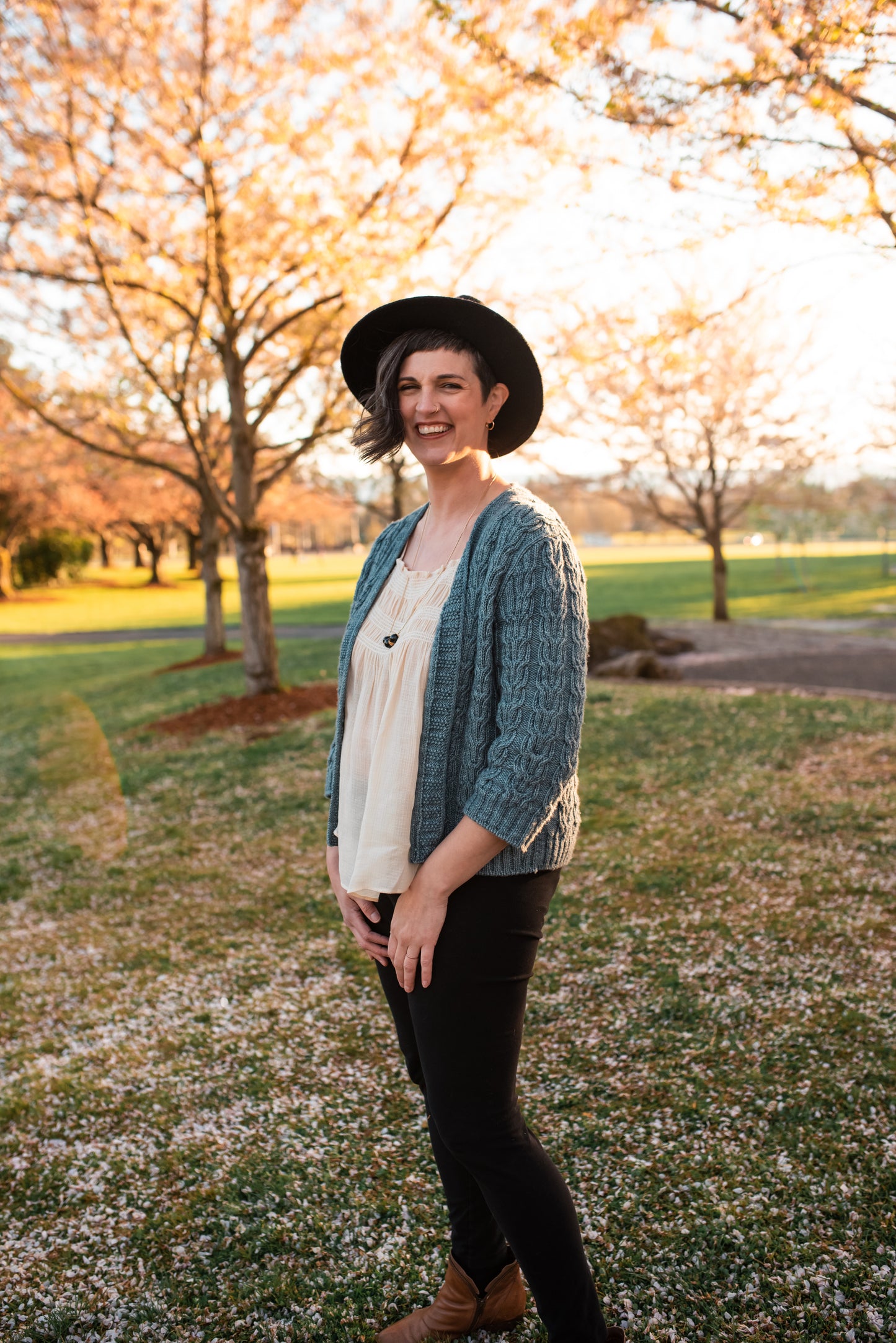 Standing in a park at a three quarter angle, Jen smiles at the camera. She wears a black hat and pants with a white tank top under a light blue cardigan. The cardigan is knit with three quarter length sleeves and horseshoe cable details.