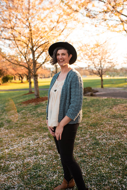 Standing in a park at a three quarter angle, Jen smiles at the camera. She wears a black hat and pants with a white tank top under a light blue cardigan. The cardigan is knit with three quarter length sleeves and horseshoe cable details.