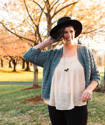 Jen grins at the camera, one hand adjusting her black hat. She wears a white tank top under a light blue cardigan knit with horseshoe cable strips.