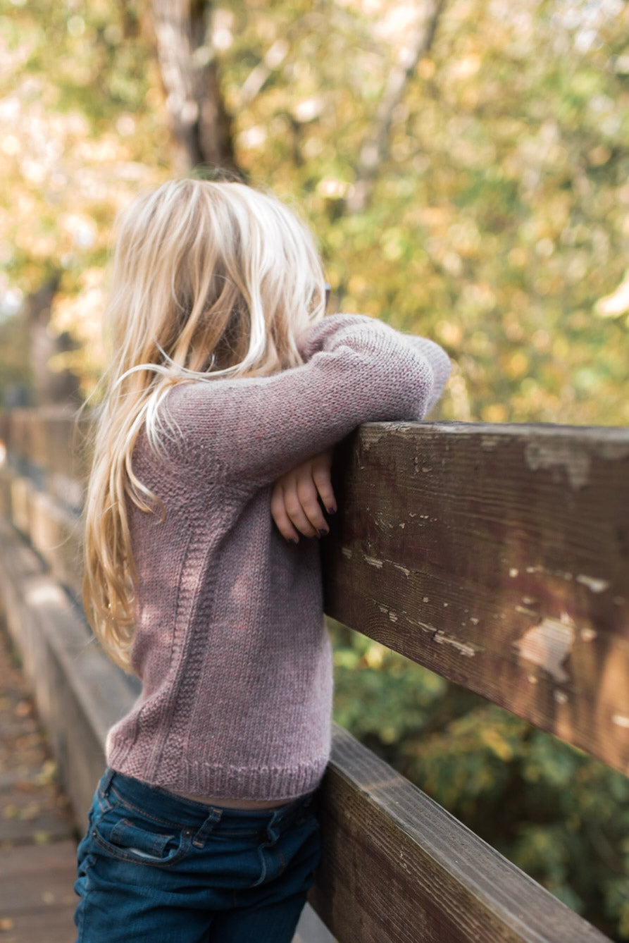 A young girl leans against a wooden railing, seen from the side. She wears a light purple sweater, knit with a subtle stitch around the seams.