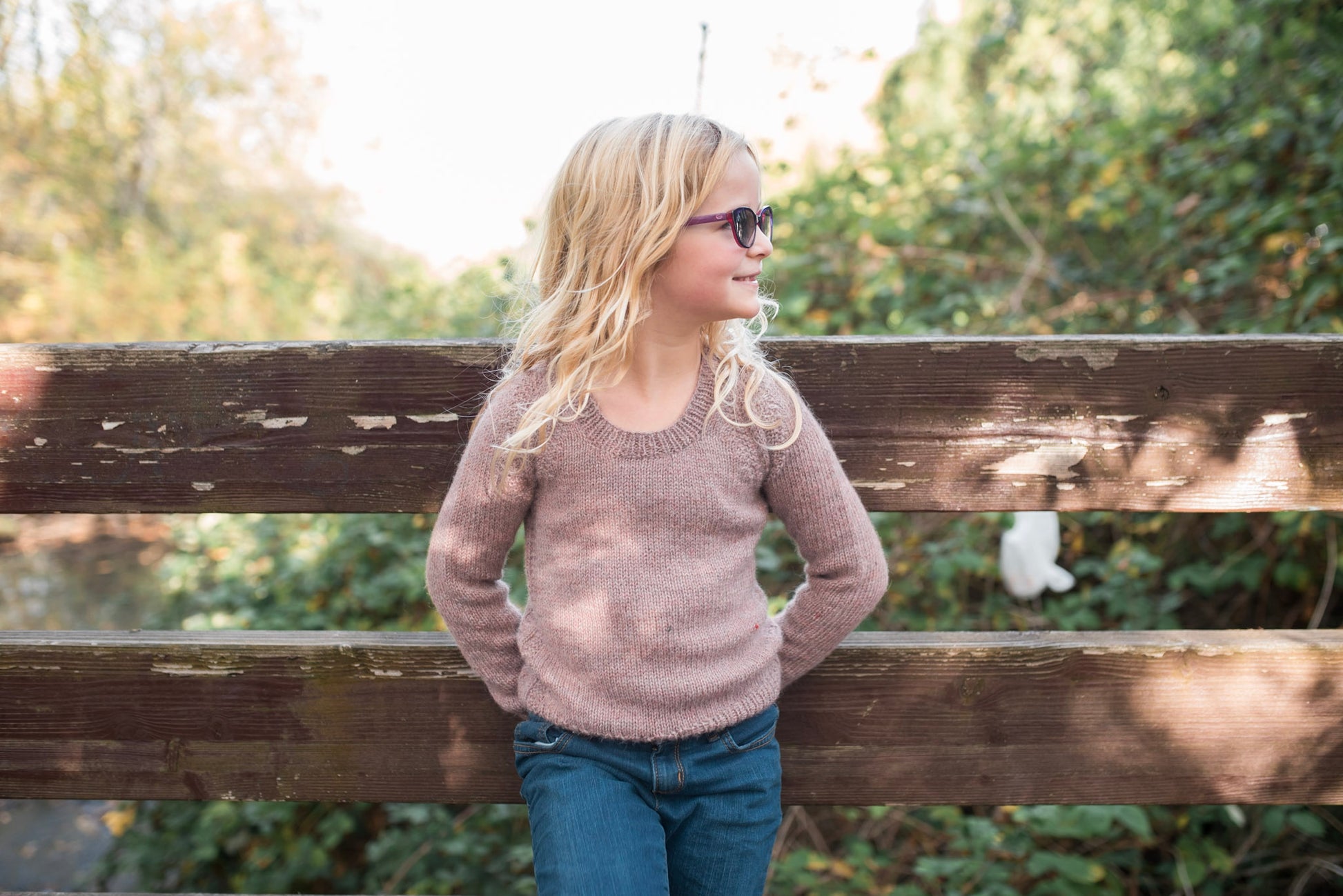 A young girl leans against a wooden railing, looking off camera. She wears a light purple sweater, knit with garter stitch embellishment around the seams and a scoop neck, with a pair of blue jeans.