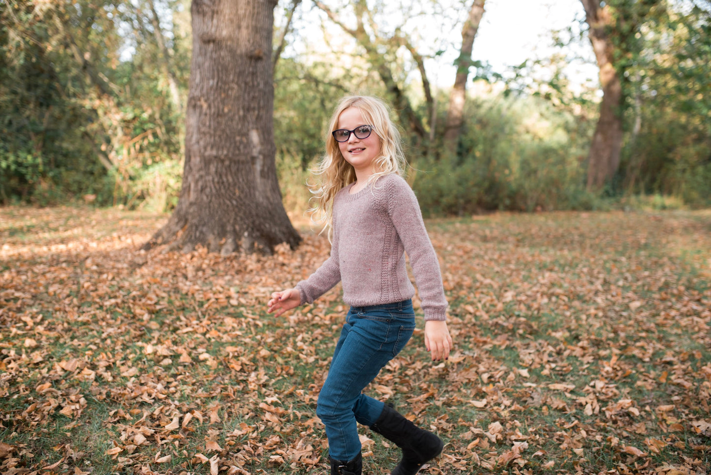 A young blonde girl walks through a park in autumn. She wears a hand knit light purple sweater, designed with a subtle garter stitch embellishment around the seams, with blue jeans and black boots.