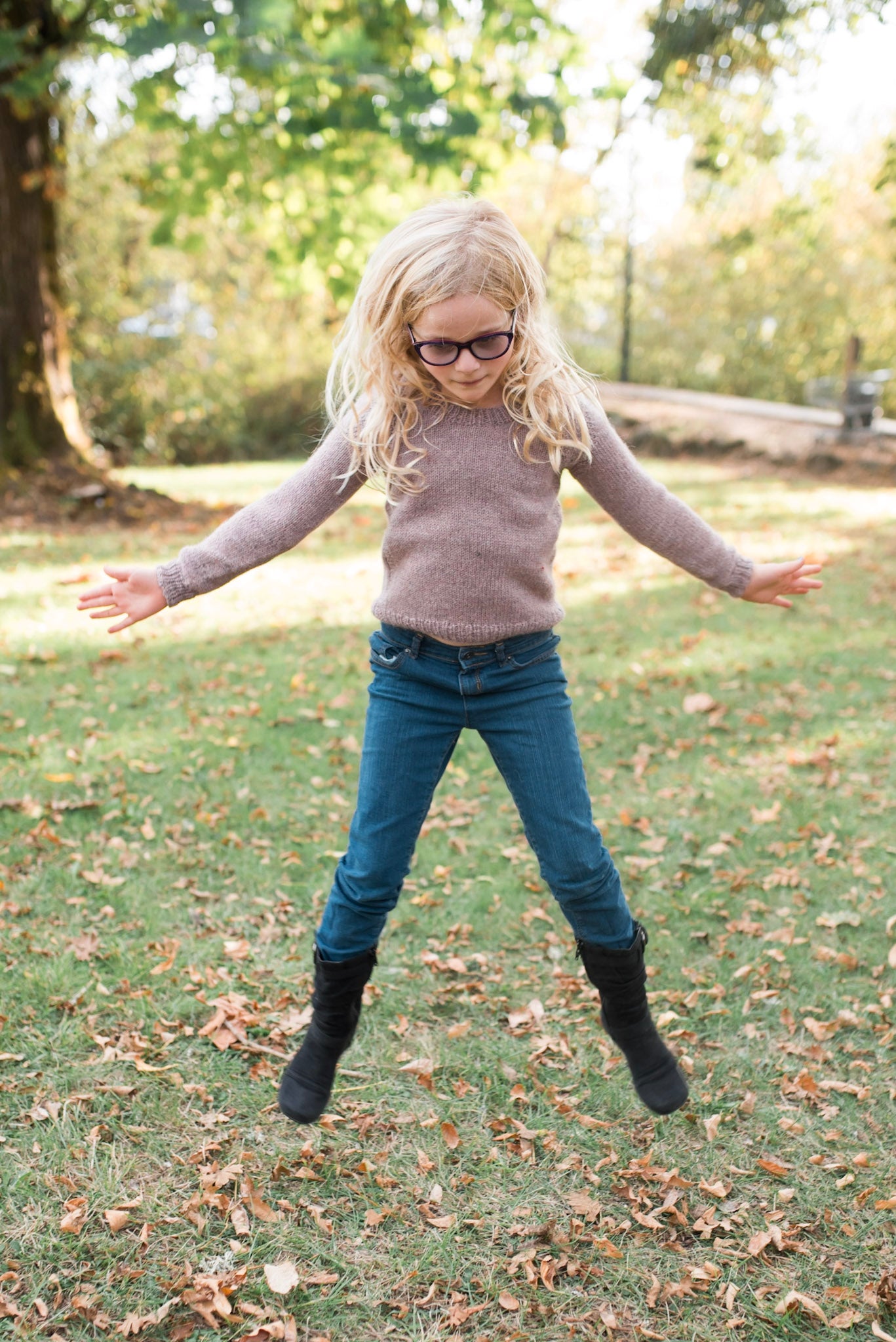 A young blonde girl, pictured jumping, wears a light purple knit pullover with blue jeans and black boots.