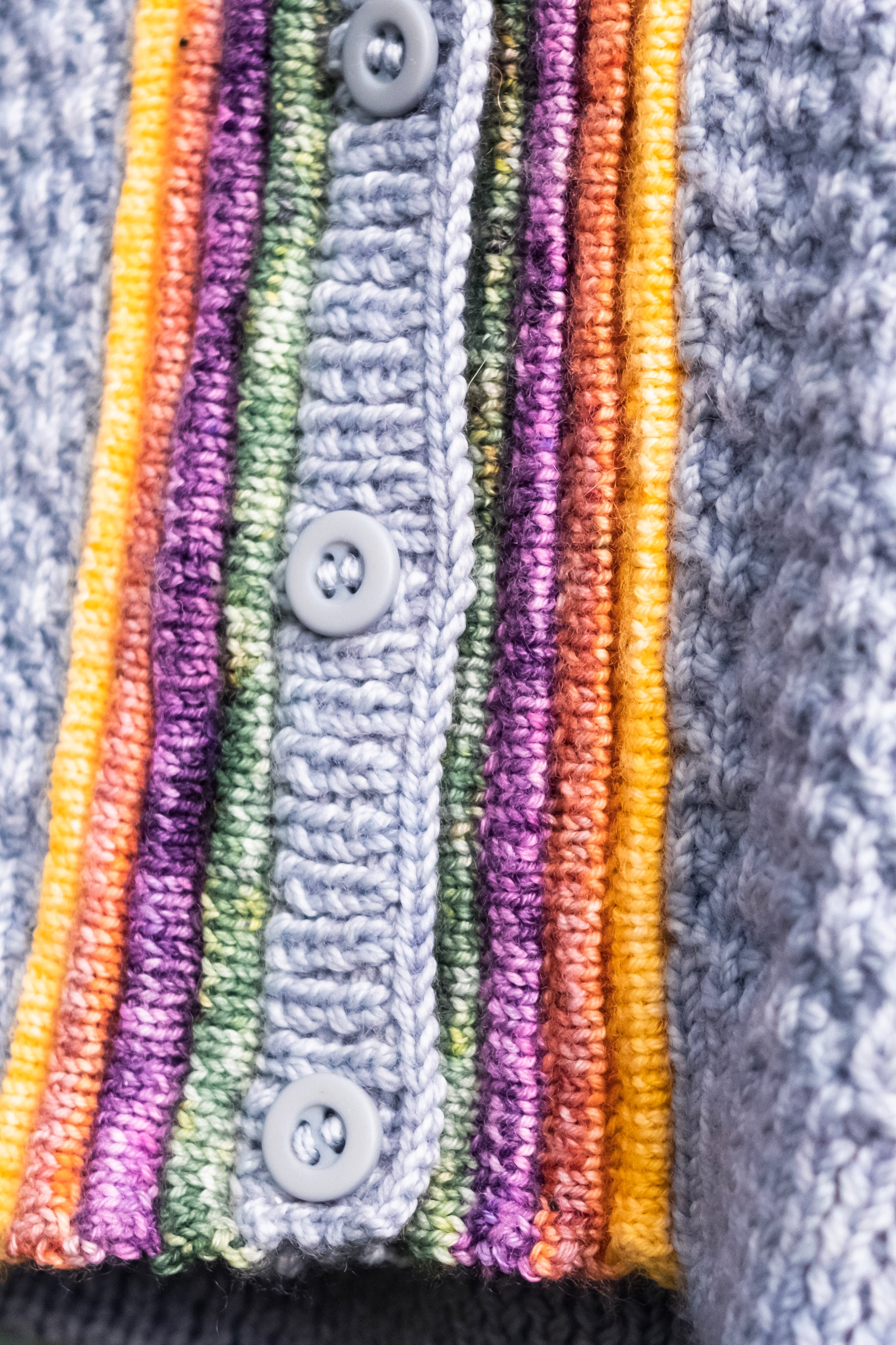 The camera focuses close up on the four plackets that surround the button band on the Letters to Camp sweater. The plackets are knit with yellow, orange, purple, and green yarn, and the sweater itself knit from blue. Three blue buttons can be seen.