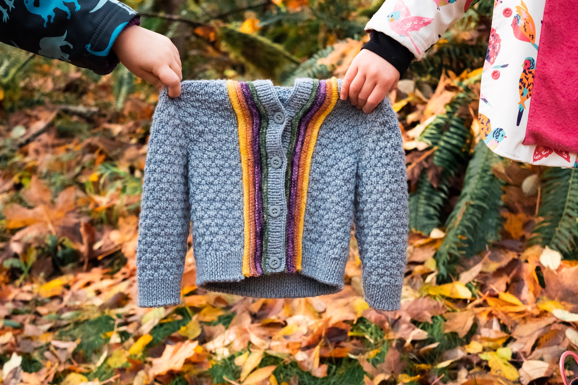 Seen close up, two kid's hold a light blue, moss stitch sweater, featuring plackets around the button band.