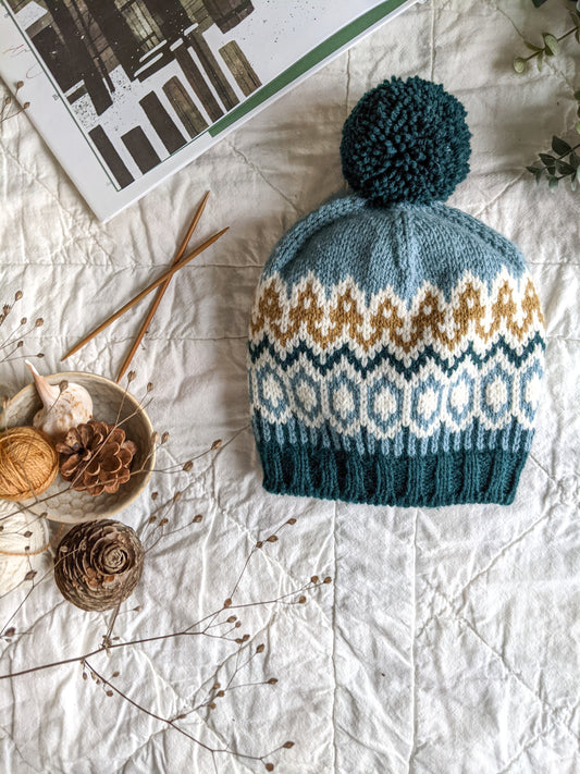 A colorwork beanie, knit with blue, orange, and white yarn and featuring a bobble, lies on a white tablecloth. Knitting needles, branches, and pine cones can be seen in the background.
