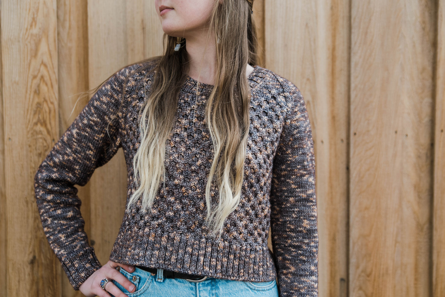 Seen from the neck down, Haley stands in front of a fence, one hand on her hip. She wears a hand knit cropped sweater, made with variegated brown yarn. A honeycomb stitch pattern adorns the body.