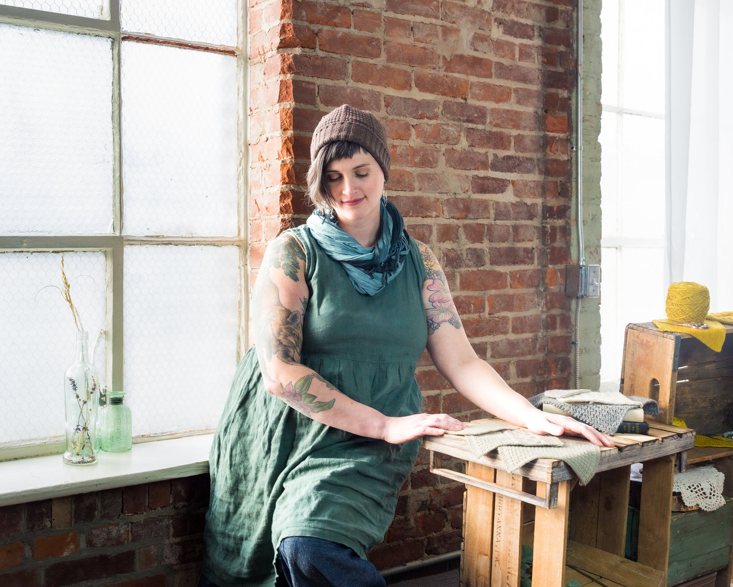 Standing against a brick wall, Jen wears a green tank dress with a brown beanie, knit with a ribbed brim and seed stitch.