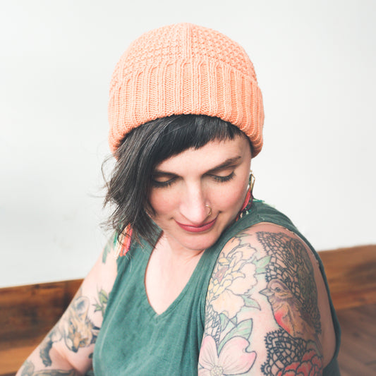 Jen, seen close up and looking down, wears a light pink hat, knit with a large ribbed edge and a seed stitch pattern, with a green tank dress.