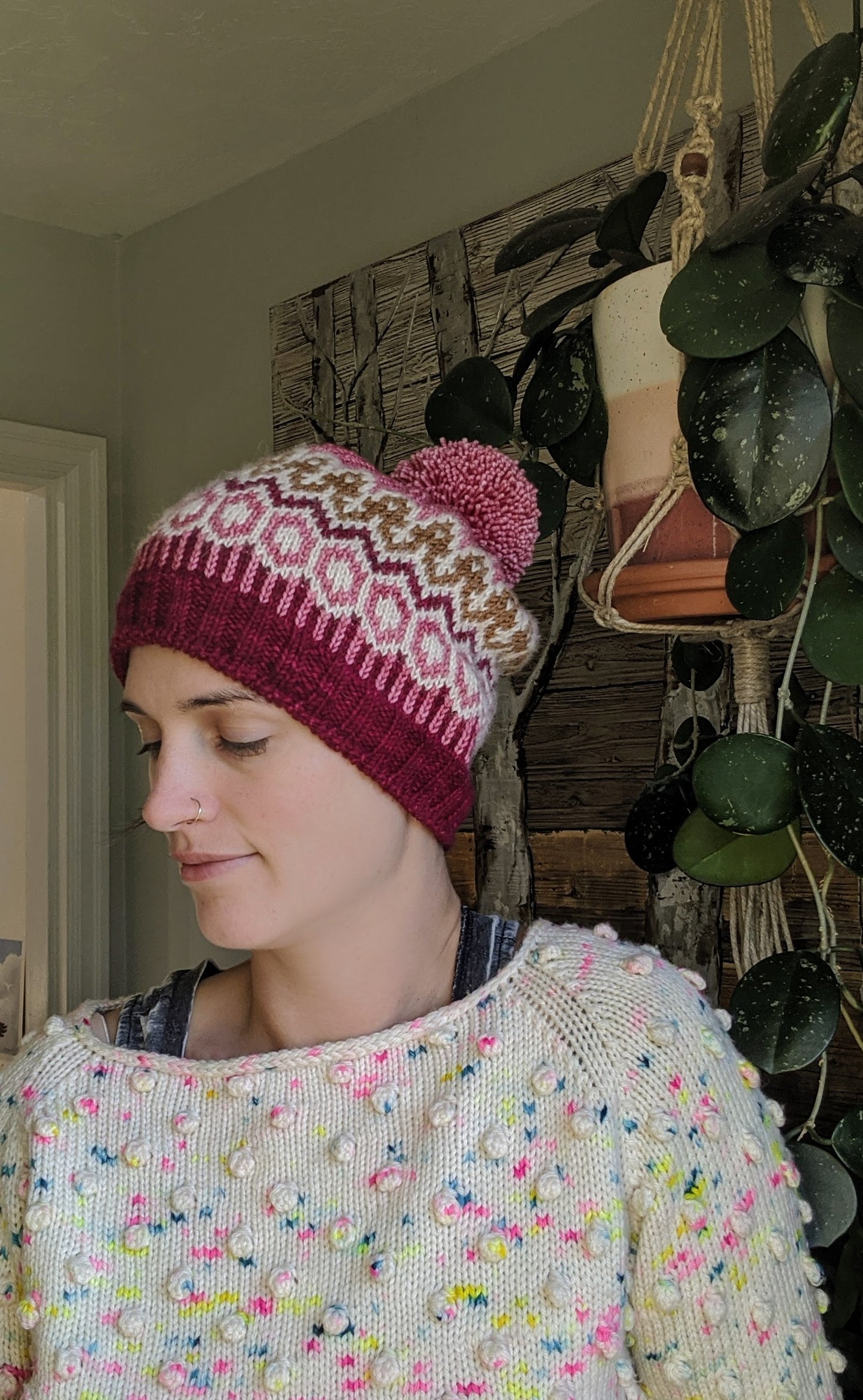 Jen wears a colorwork knit hat, made from dark pink, brown, white, light pink yarn with a bobble on top. Houseplants can be seen in the background.
