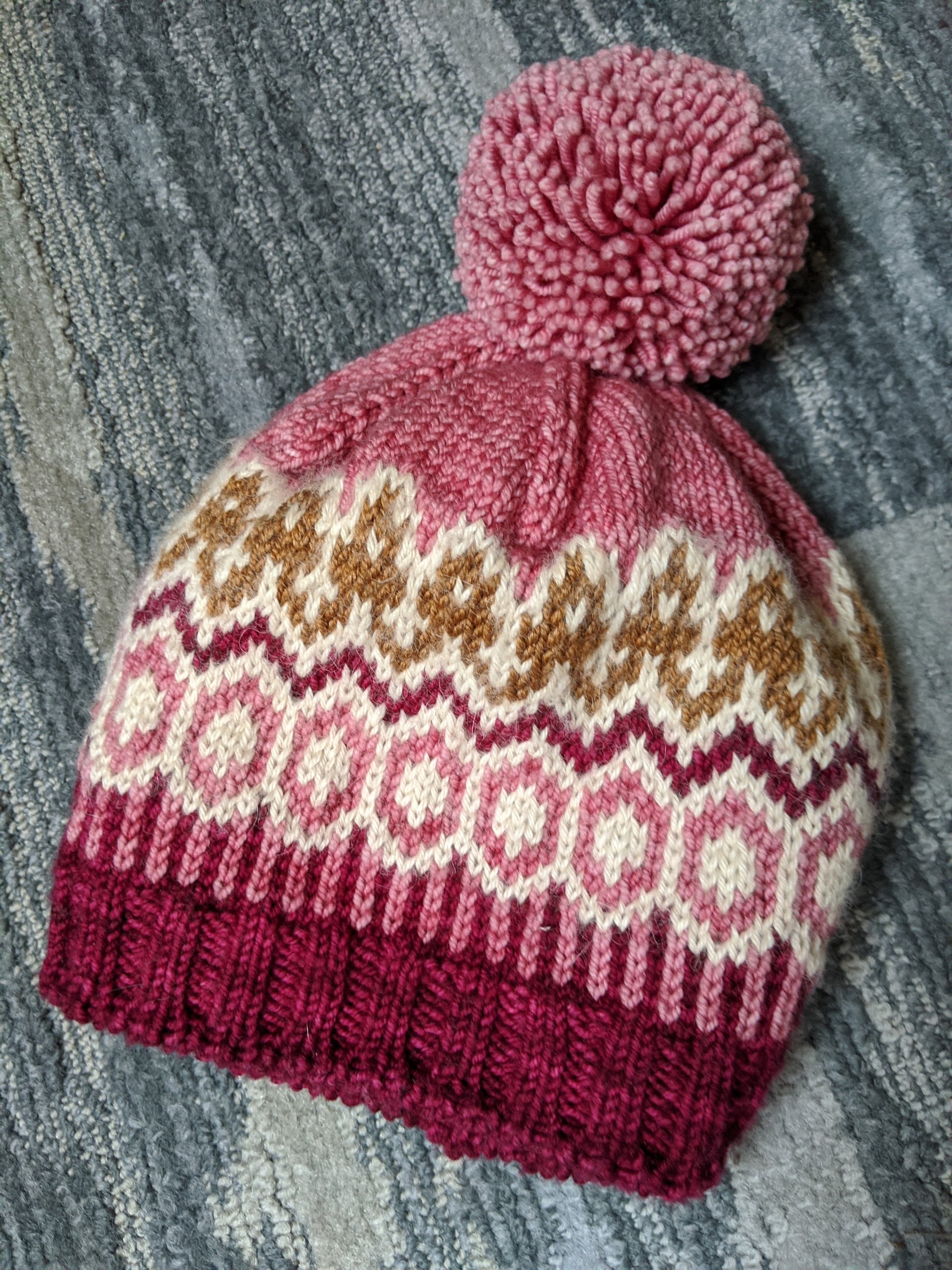 A knit beanie, made from light pink, white, brown, and dark pink hat, lays on a rug. It's made with a yarn bobble on top.