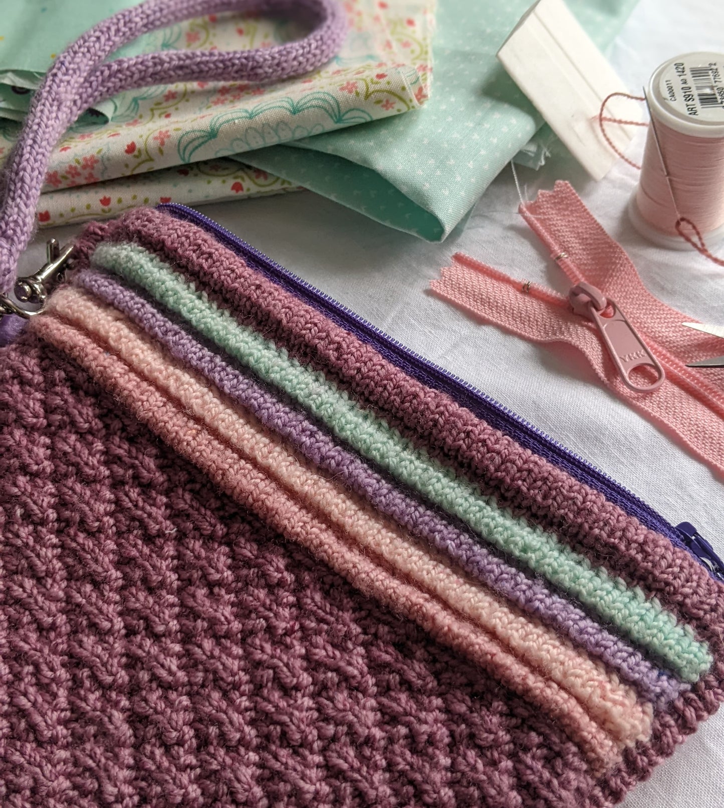 A Ziplet pouch, knit in dark pink, orange, green, and purple yarn lays on a table. Blue fabric, a pink zipper, and a needle and thread can be seen in the background.