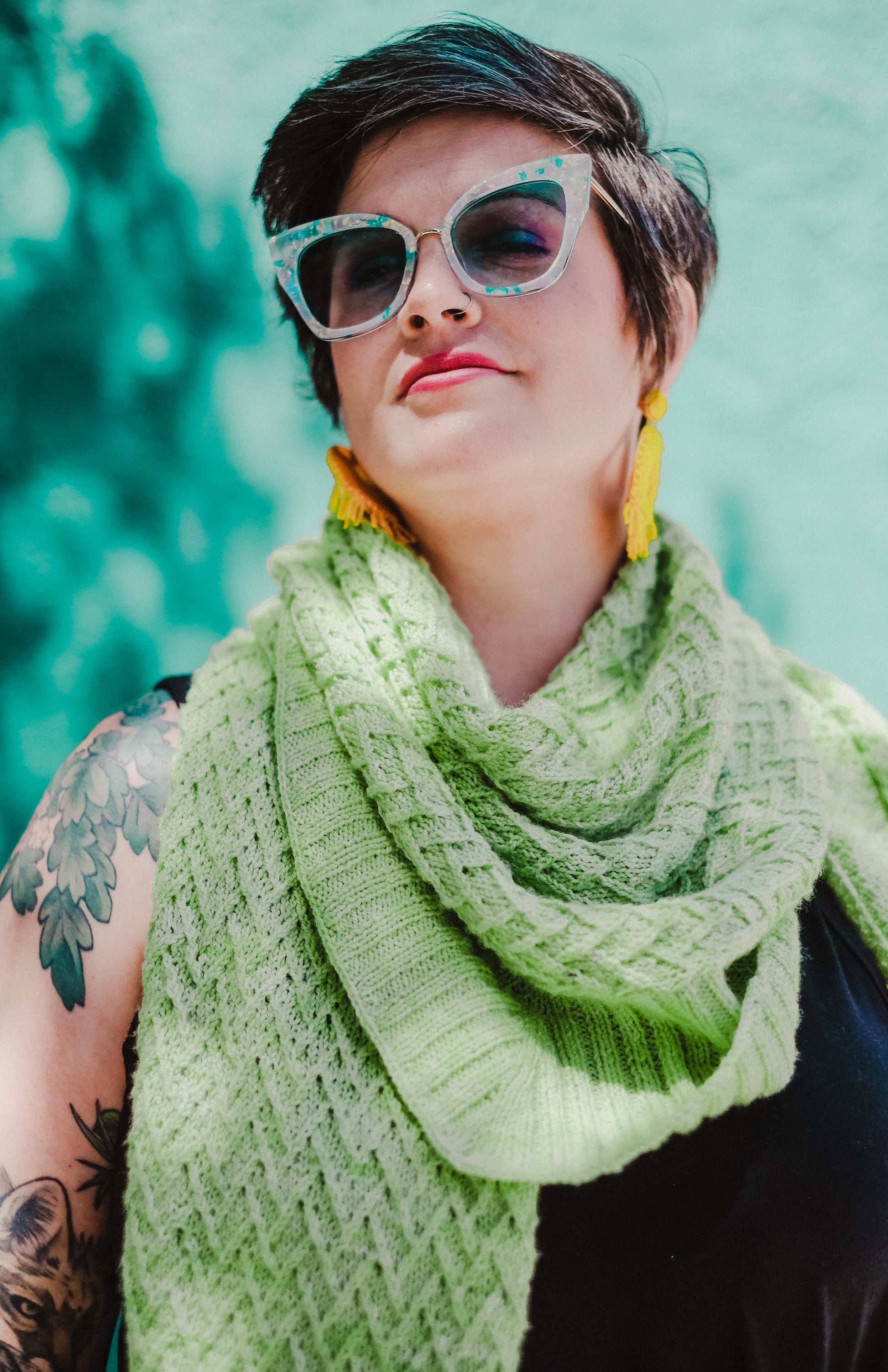 Jen, head tilted towards the sun, wears a hand knit, lime green shawl with sunglasses and large tassel earrings. The shawl has a chevron cable pattern with a ribbed edge.