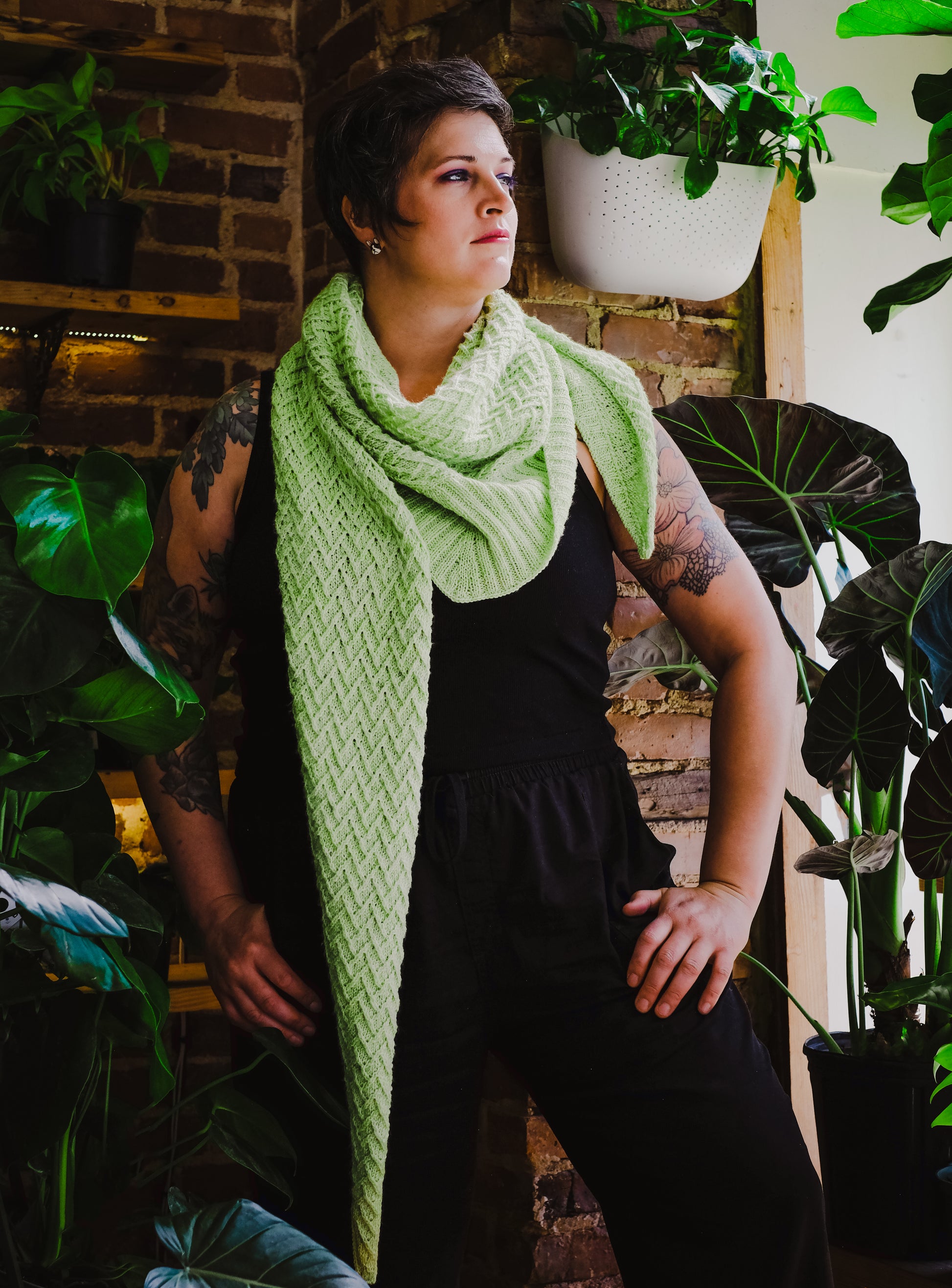 Jen stands, hands on her hips, looking off camera. She wears a black tank and pants under a lime green shawl, knit with chevron details in an asymmetric triangle.
