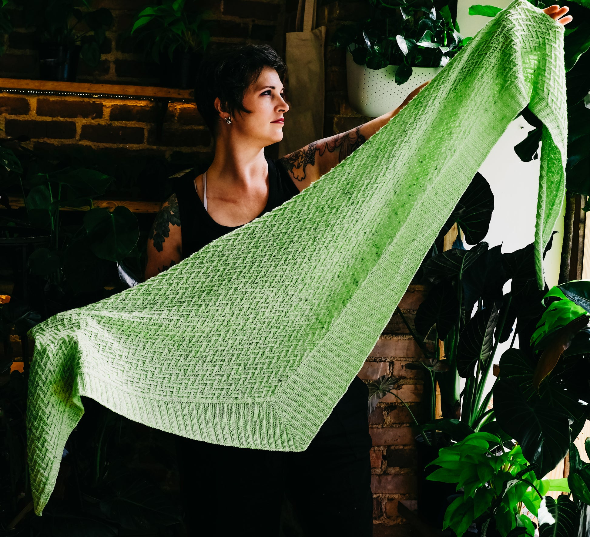 Seen from the front, Jen looks off camera, holding open a triangle, hand knit shawl. Knit in lime green yarn, the shawl features a chevron pattern and a ribbed edge along two sides of the triangle.