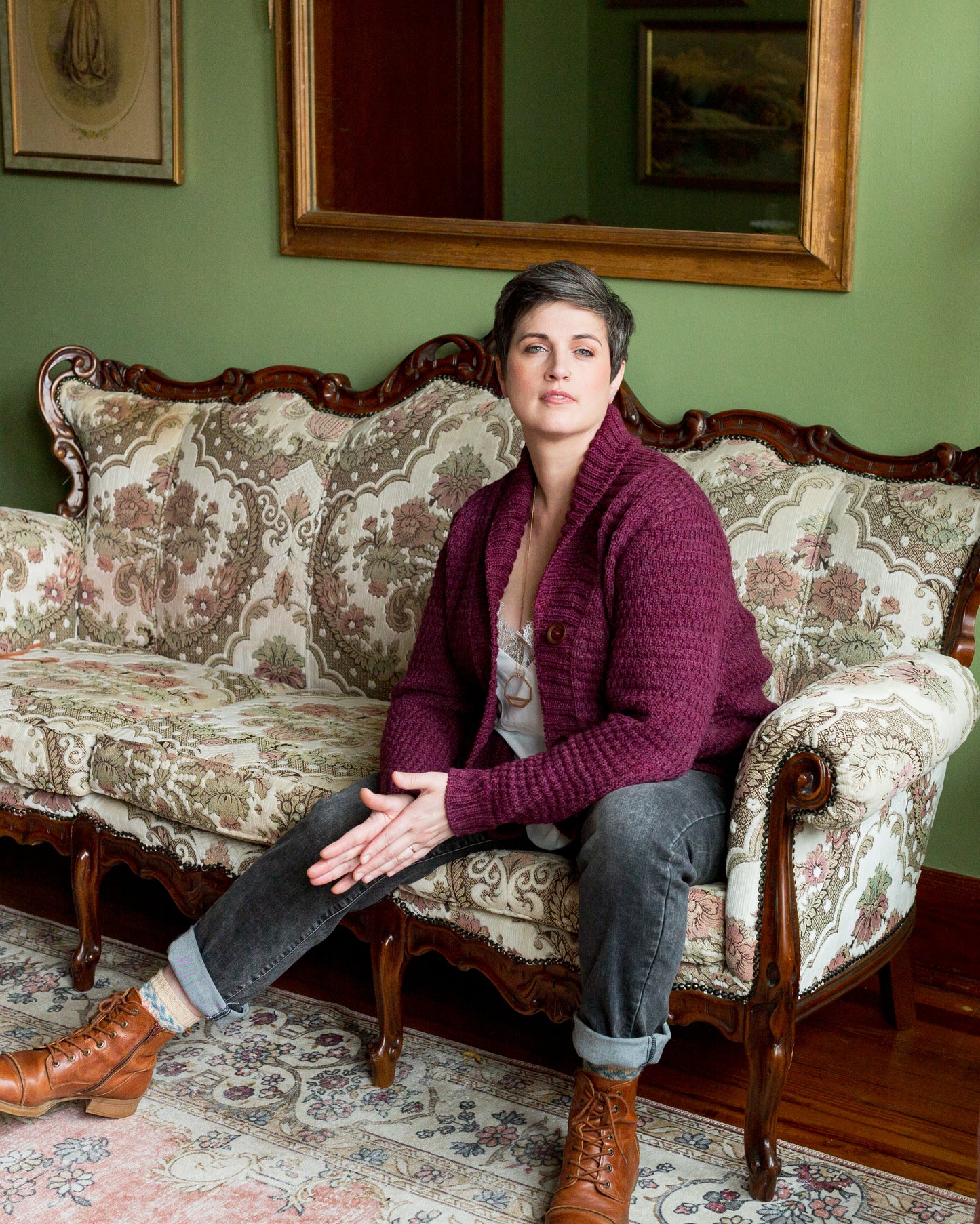 Jen sits on an antique couch, leaning forward slightly to look in the camera. She wears a burgundy cardigan with brown boots and grey jeans. The cardigan is knit with a cowl neck and a subtle all-over stitch pattern.