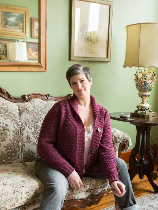 Jen sits on an antique couch, looking at the camera. She wears a cowl neck cardigan, knit with moss stitch from a burgundy yarn. An antique lamp and painting can be seen in the background.