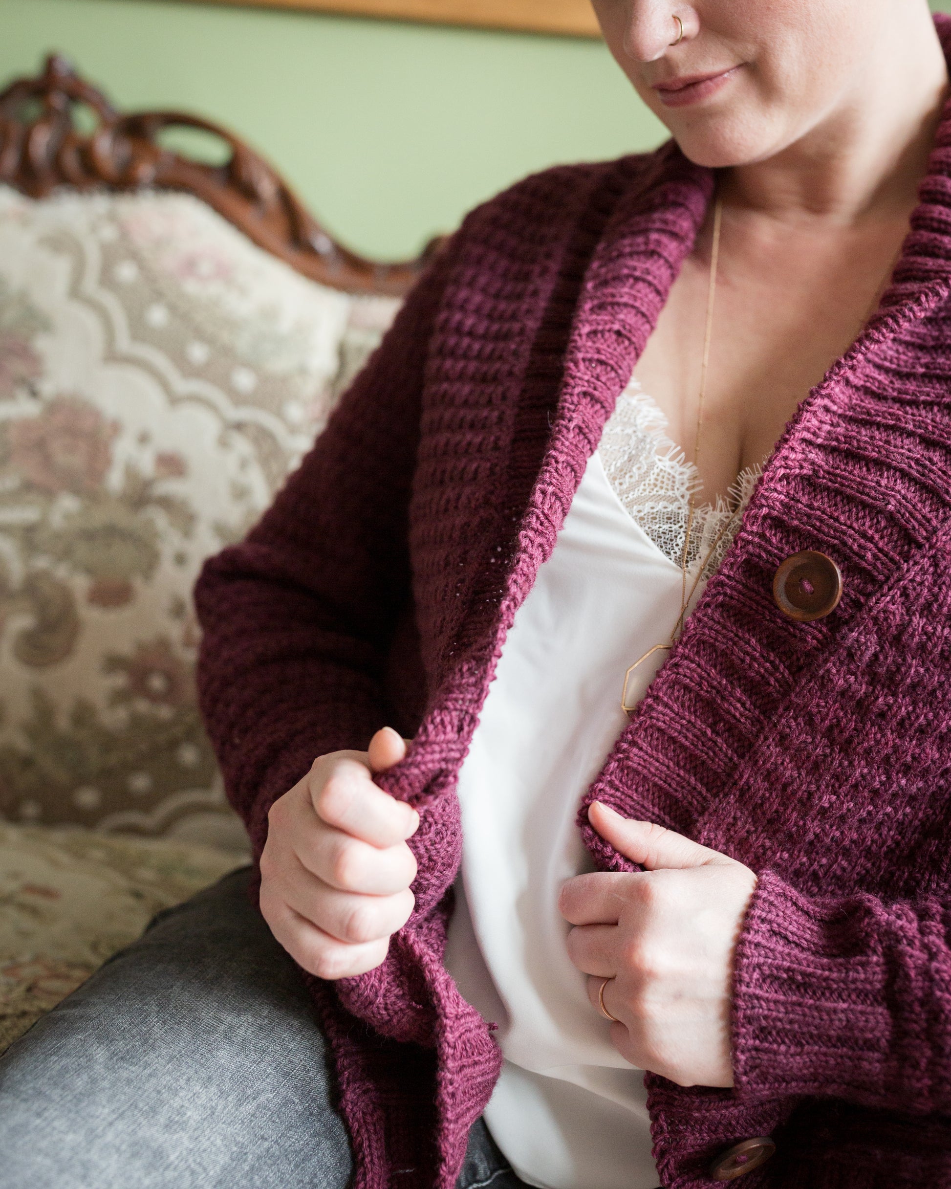Seen from the shoulder down, Jen pulls the sides of a burgundy cardigan together, showing off the hand knit details of the large button band and subtle stitch pattern.